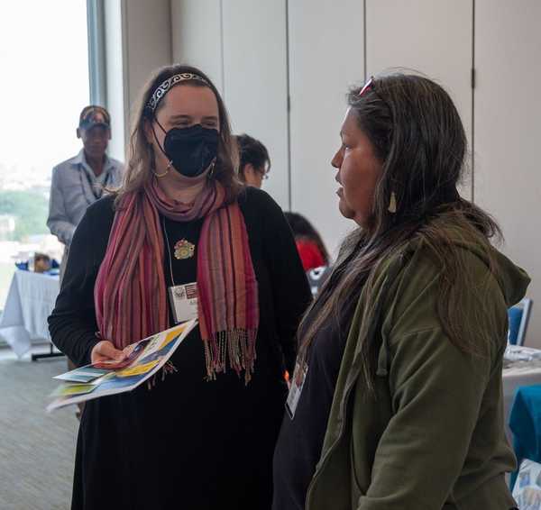 Allison Taylor, a white woman on left wearing a black mask, black dress, and colorful scarf, talks with Ella Blackowl, an Indigenous woman in a green coat, also from SGISD.